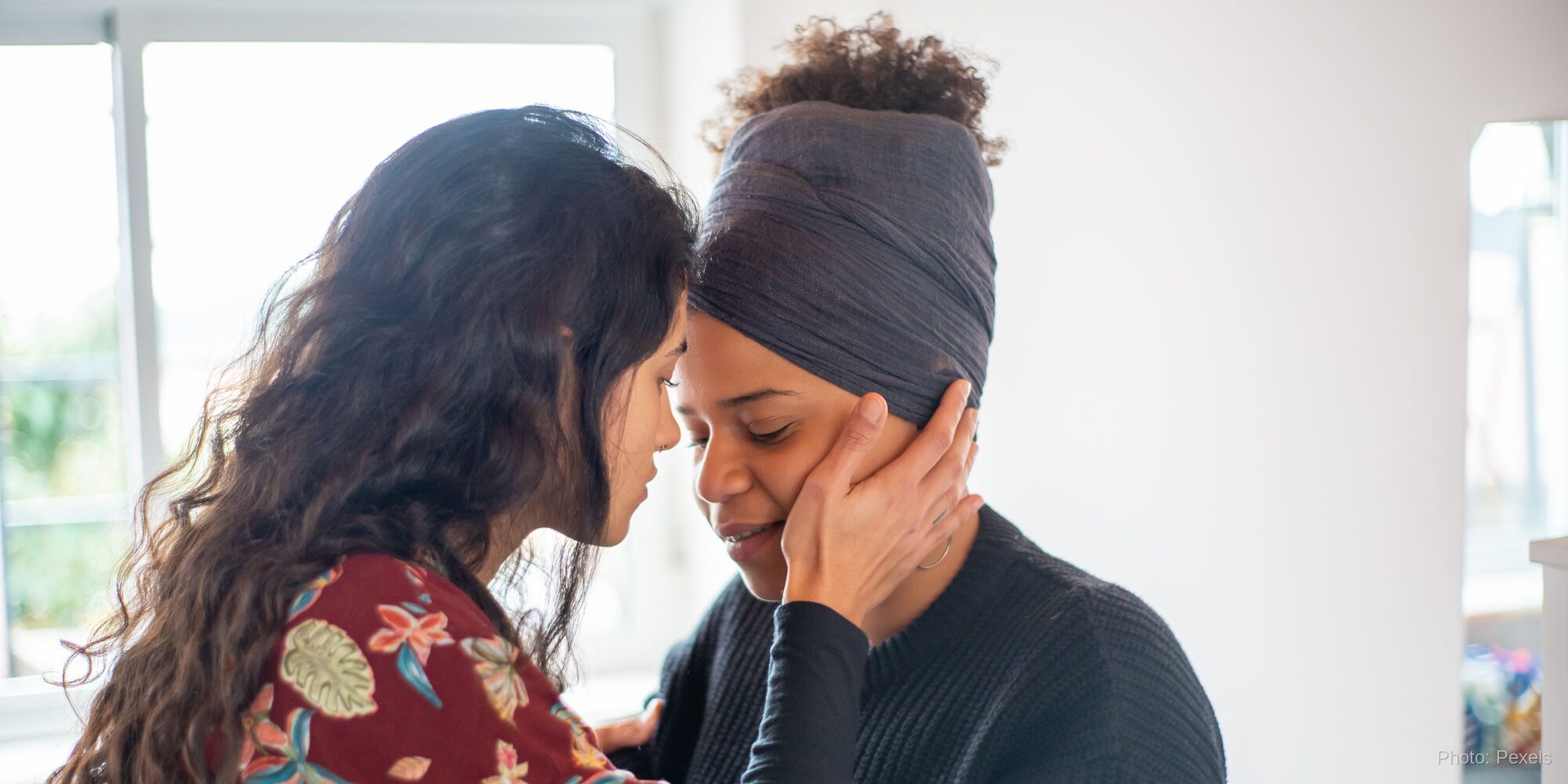 One woman gently cradles another's face in her hands, foreheads close, in a moment of tender, wordless comfort.