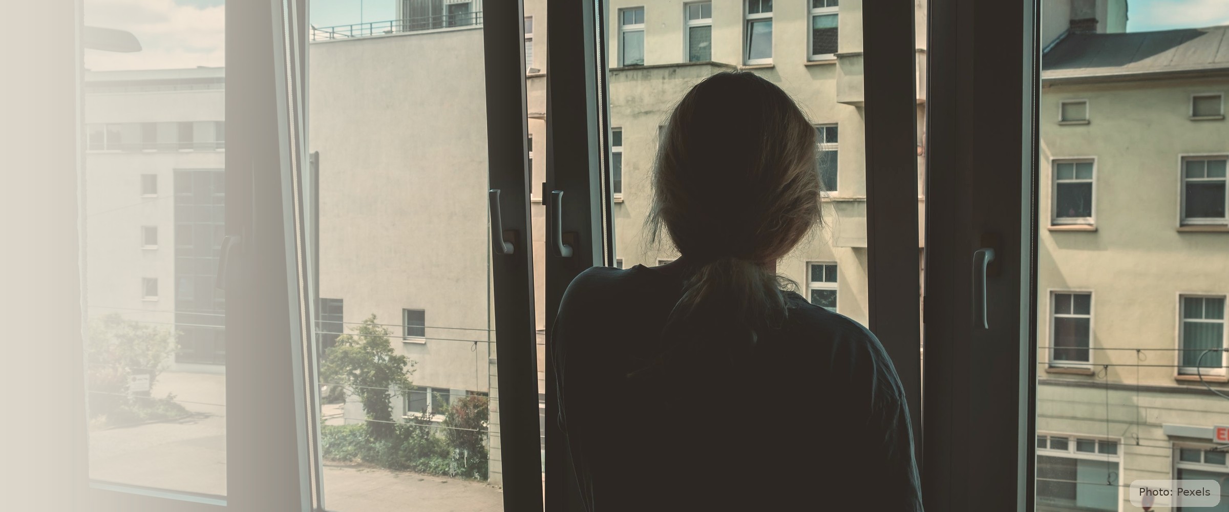A woman, seen from behind, stands quietly looking out an open window — the after, not the violence.