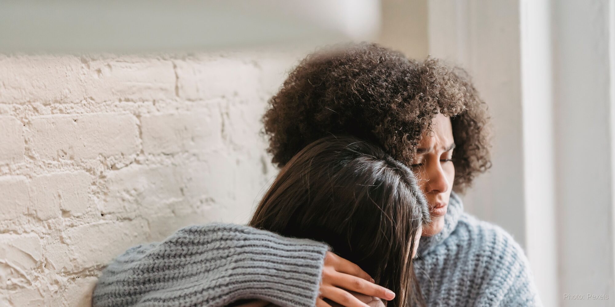 Two women embrace closely, one resting her head on the other's shoulder in a quiet moment of comfort and care.
