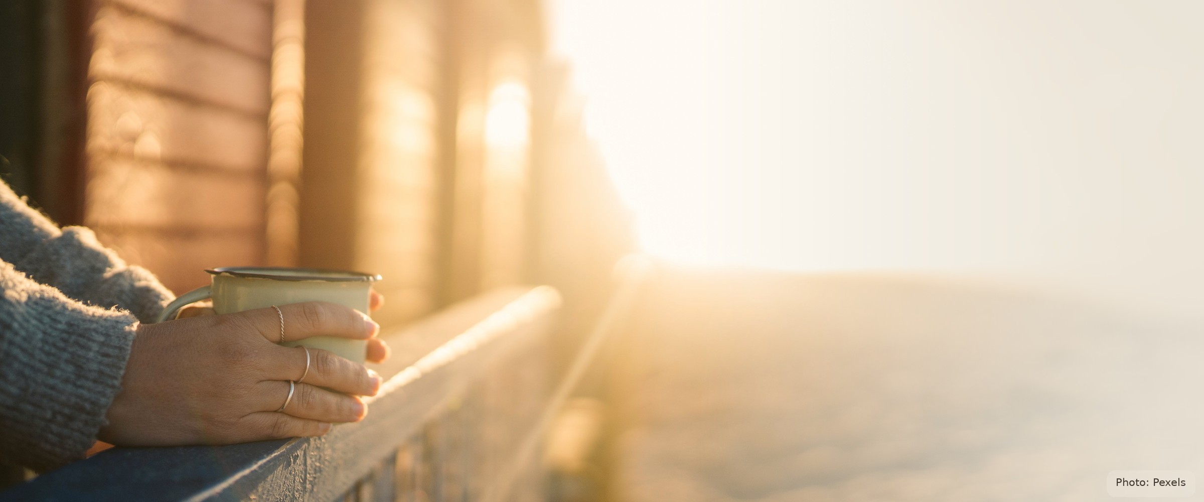 A hand wrapped around a warm mug rests on a porch railing at sunrise — steadiness, not the crisis.