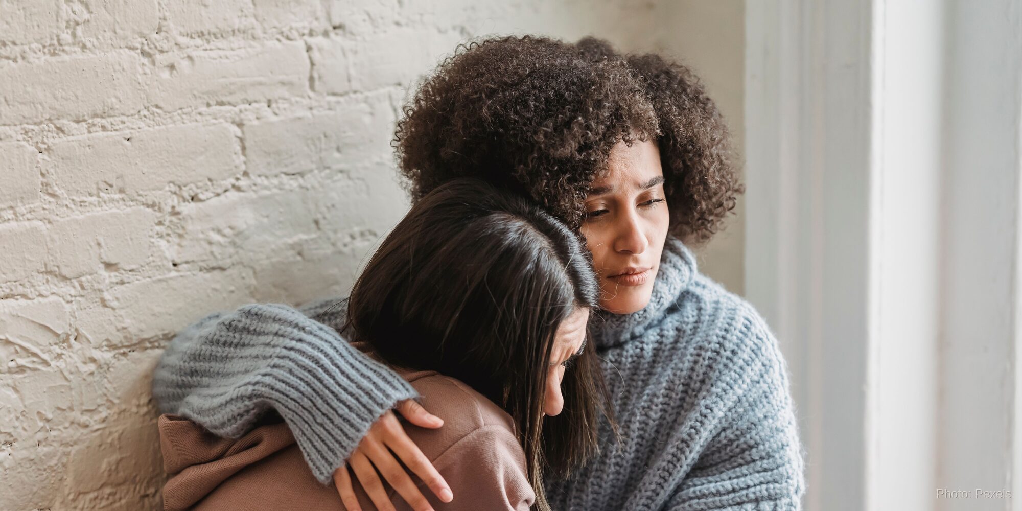 Two women share a close, comforting embrace, one gently resting a hand on the other's head.