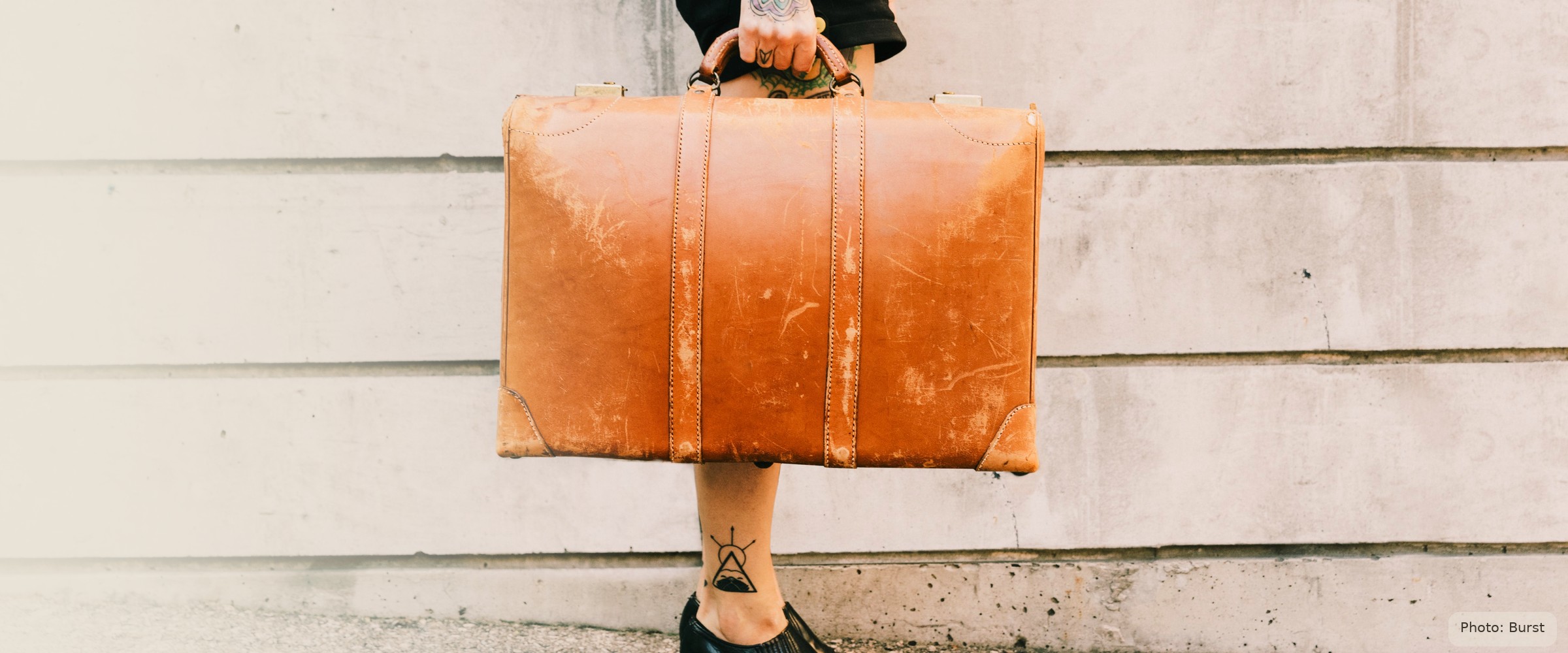 A young woman's hand carries a worn vintage suitcase against a plain limestone wall — starting adult life with what she can carry.