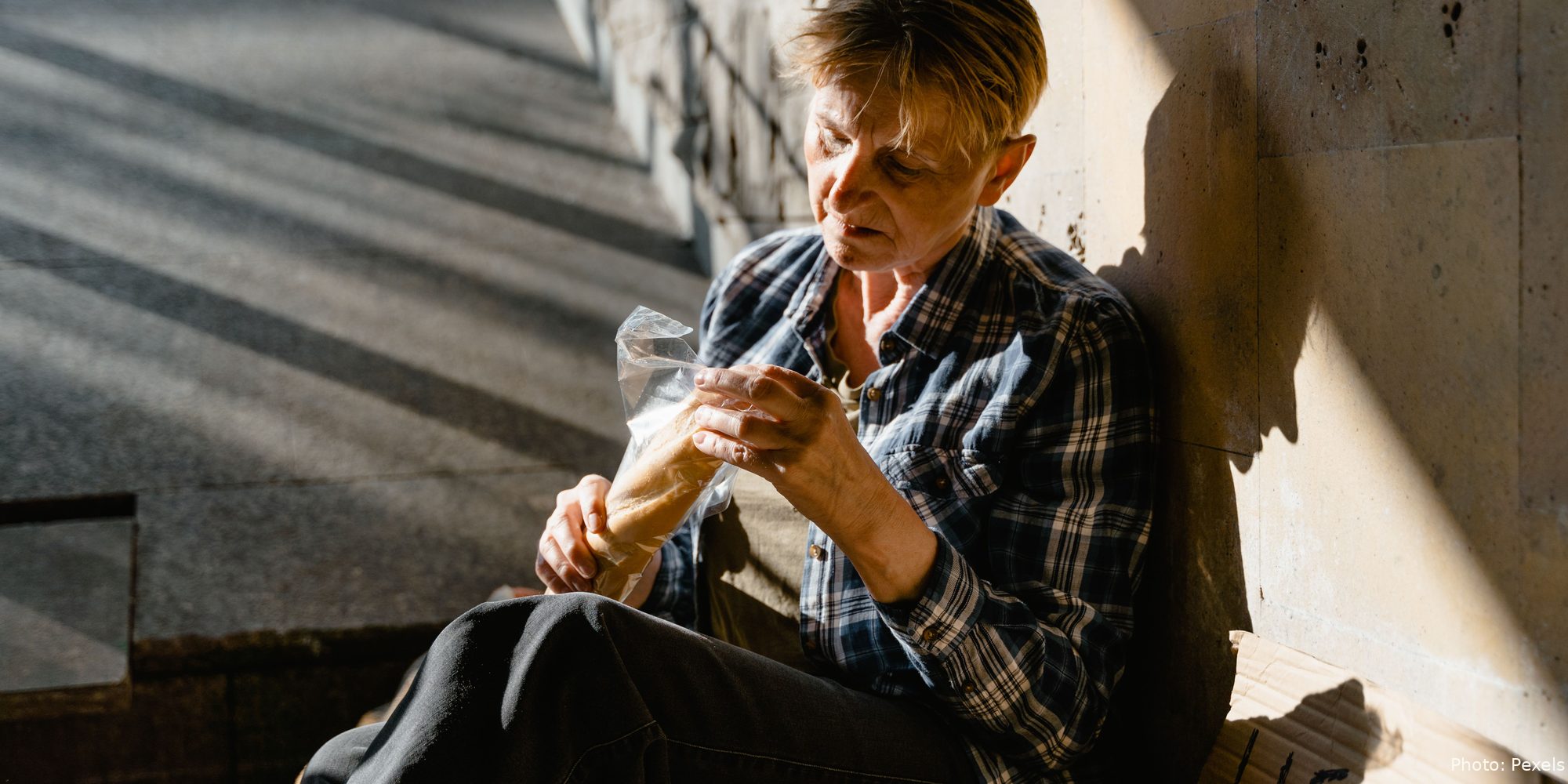 An older woman sits on stone steps in a patch of morning sun, holding a small loaf of bread.