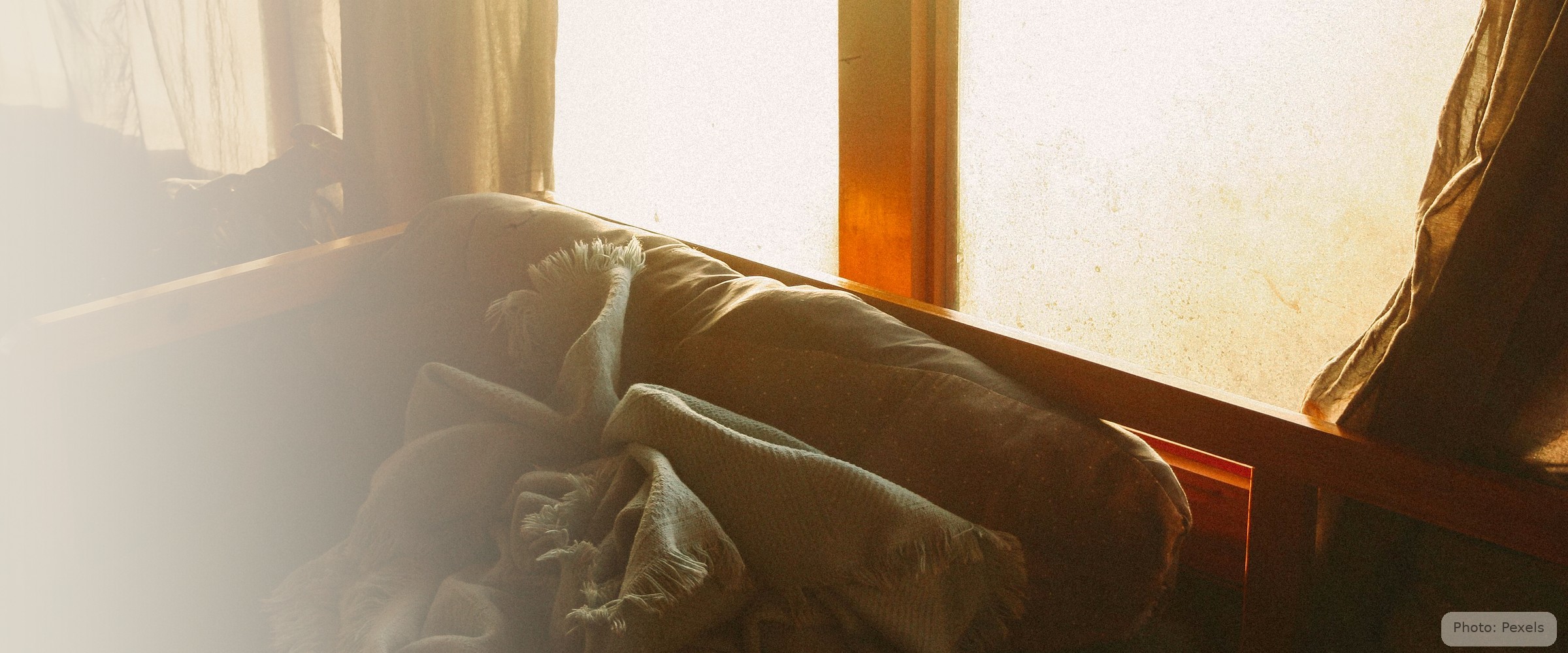 A folded blanket rests on a worn sofa cushion, warm window light behind — the hidden kind of homelessness.