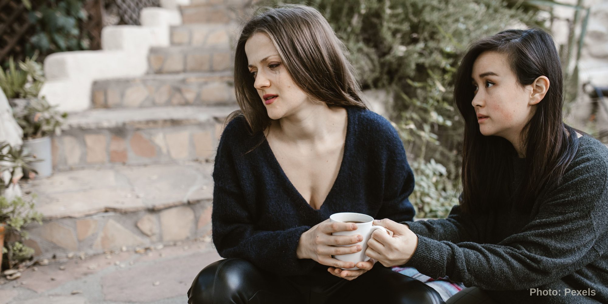 Two women sit talking on outdoor stone steps, one holding a mug, the other listening with a hand of support nearby.