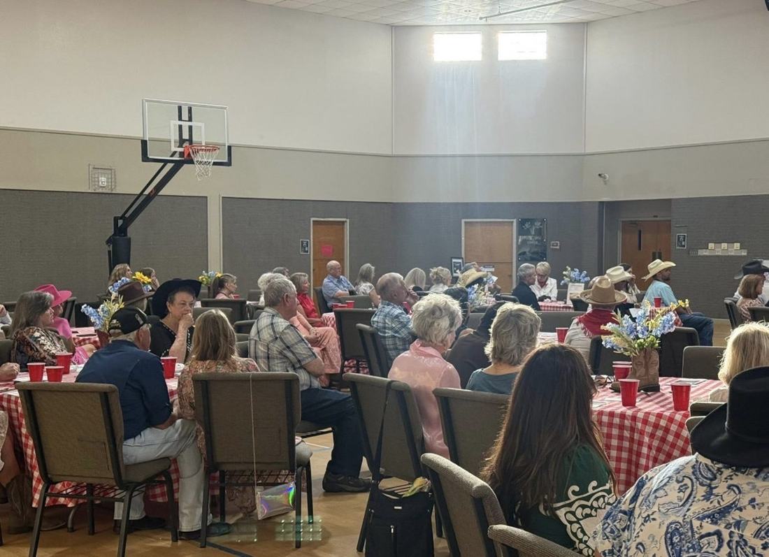 A church hall filled with guests seated at tables covered in red-and-white gingham, listening to a speaker.