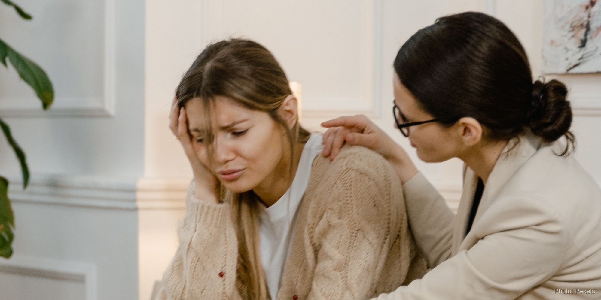A distressed woman holds her head while a trusted friend in professional dress rests a hand on her shoulder in support.