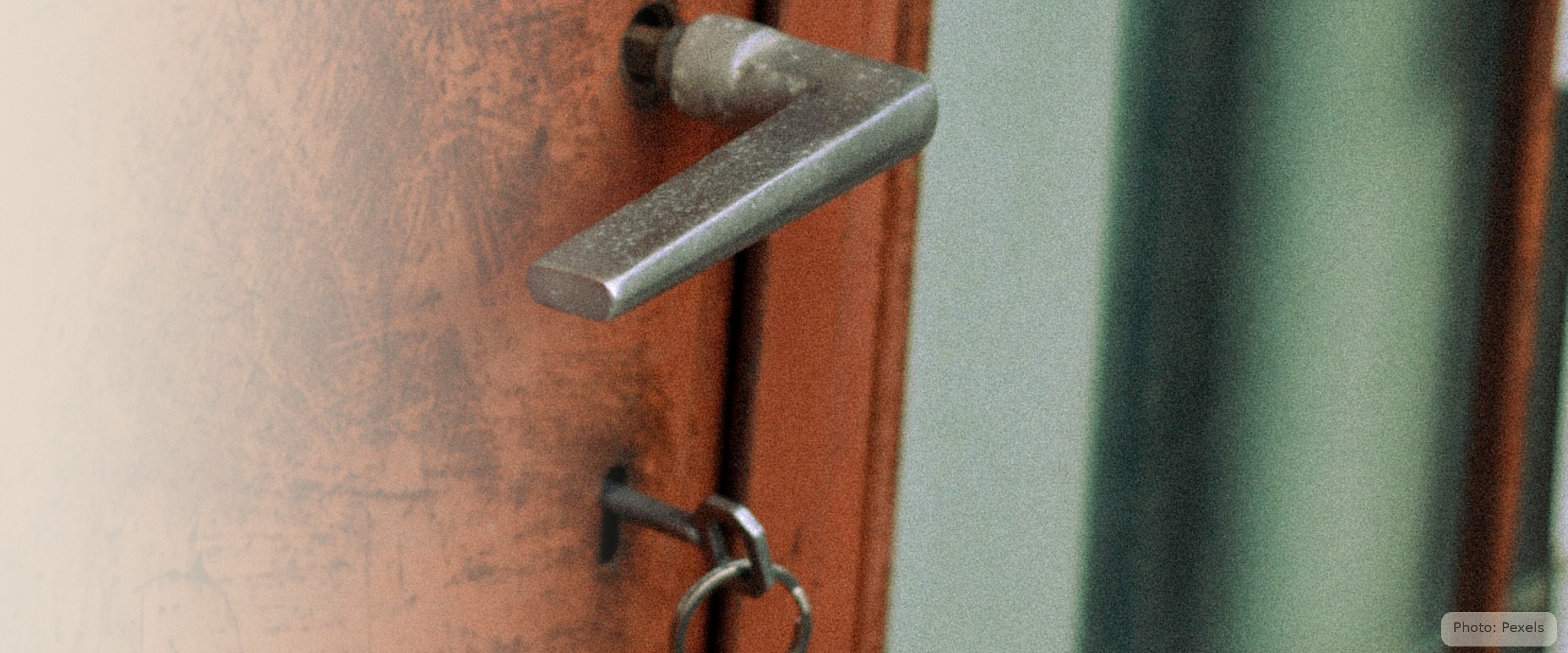 A set of keys hangs in the lock of a weathered clay-red door, morning light beyond.