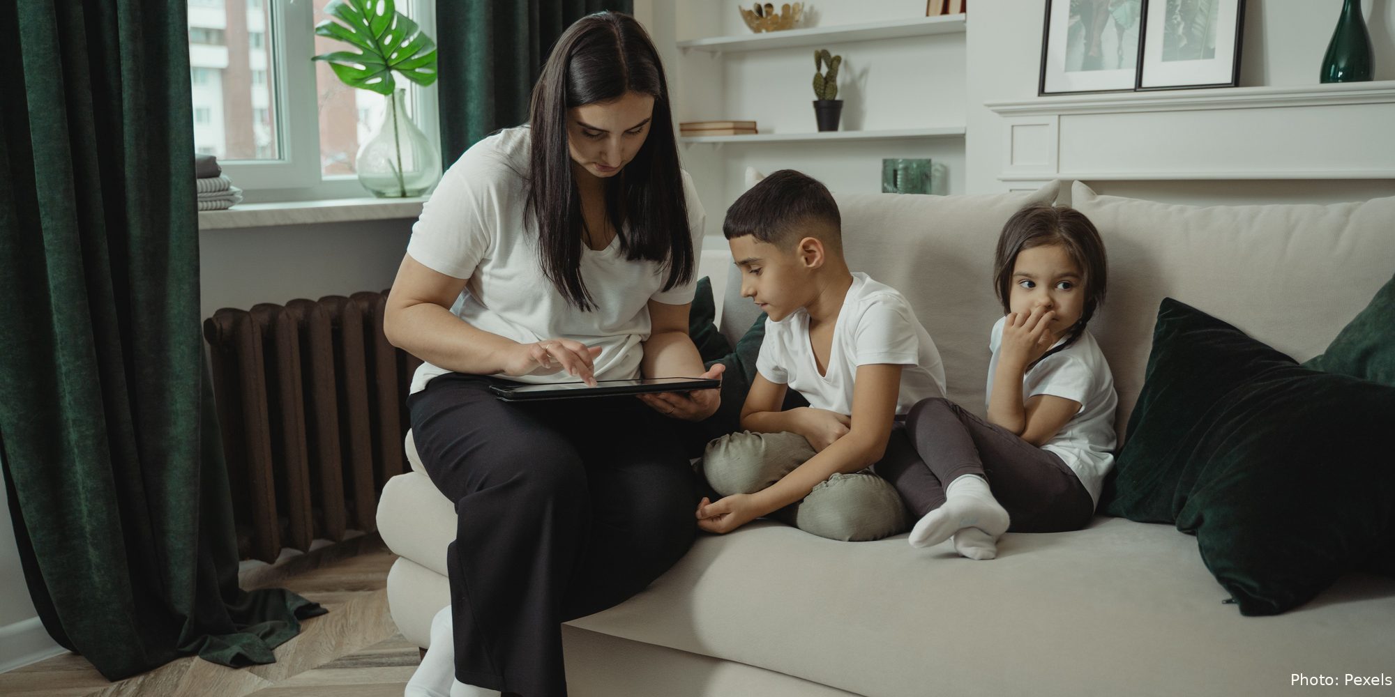 A mother and her two children sit close together on the couch, looking at a tablet.