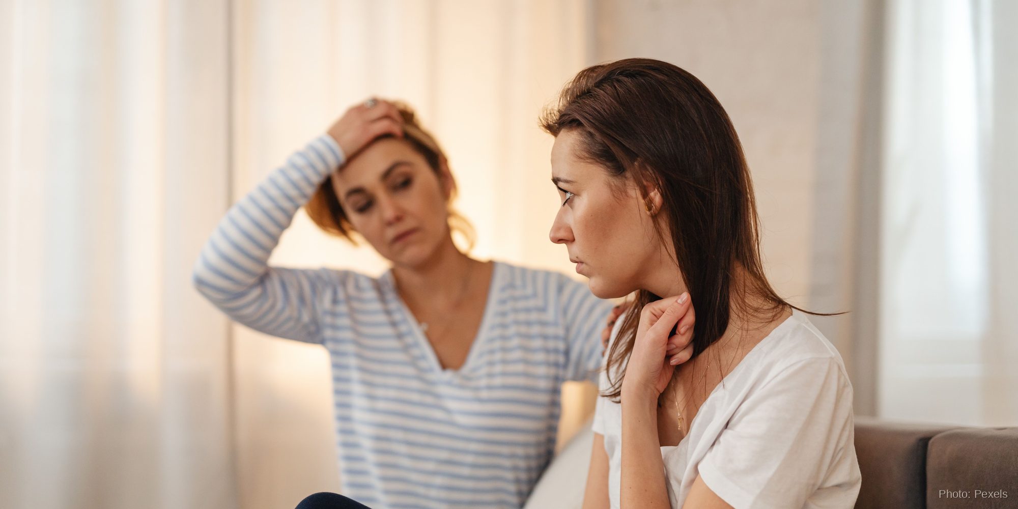 Two women sit close in quiet conversation, one listening with visible concern as the other looks down in thought.
