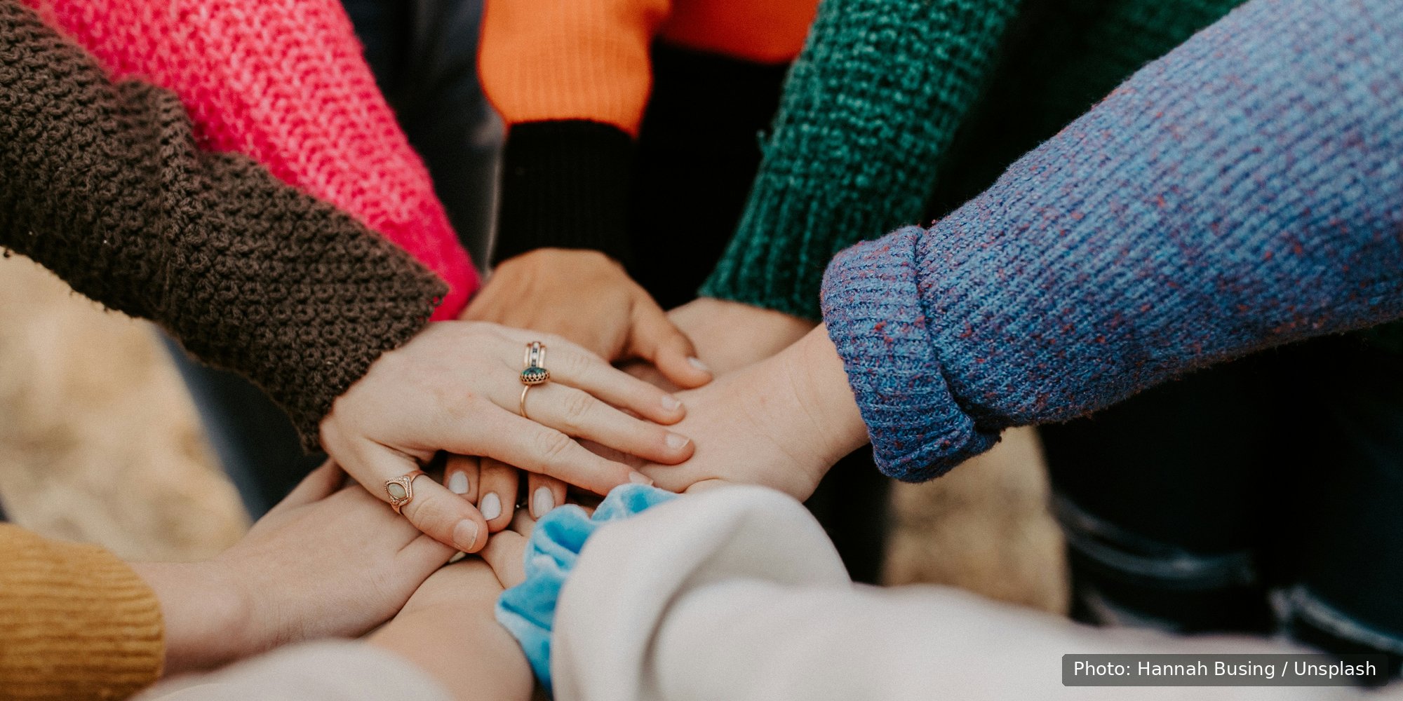 Hands from several people in colorful sweaters stack together in a circle of support.