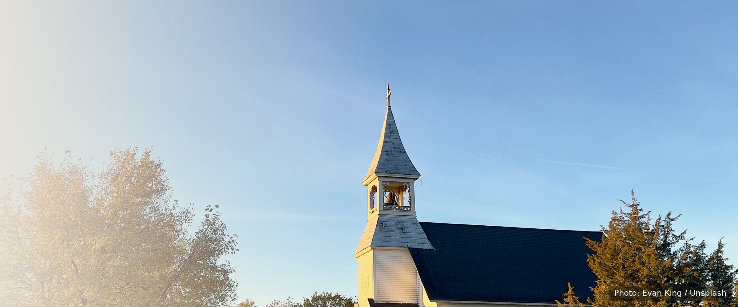 A small white church with a black roof and steeple stands against a clear blue sky at golden hour.
